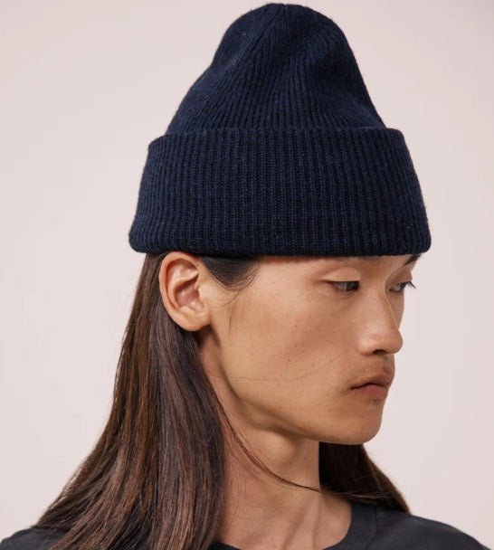 The HARTFORD - SEAMLESS WOOL BEANIE IN NAVY by HARTFORD is a dark blue ribbed knit beanie made from 100% wool, featuring a folded brim and shown against a plain light gray background.