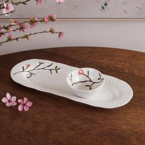 A white BEATRIZ BALL CERAMIC BIRD ON BRANCH MINI BOWL sits on a wooden table, accented by hand-painted cherry blossom branches, pink blossoms, and a decorative branch.