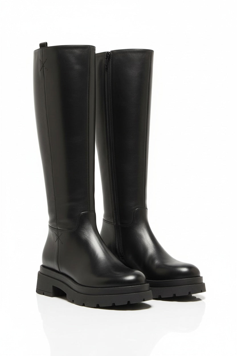 A person stands against a plain white background wearing the TALL LEATHER BOOT by MICHELE LOPRIORE—crafted from genuine leather, featuring chunky soles, and made in Italy.
