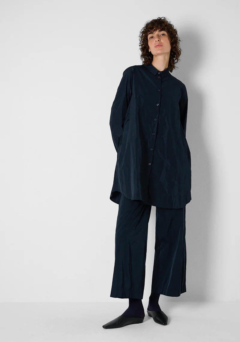 Against a light gray background, a person models the KATHARINA HOVMAN - CLASSIC OVERSIZED SHIRT with mother-of-pearl buttons and black pants. Their hair is tied back as they face away, slightly raising their arms to highlight the shirt's loose fit.