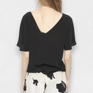 A woman with long blonde hair is wearing a black, short-sleeved MEIMEIJ - Flutter Sleeve Blouse and white pants with a black floral pattern. She is standing against a plain, light-colored background. Her face is not visible in the image.