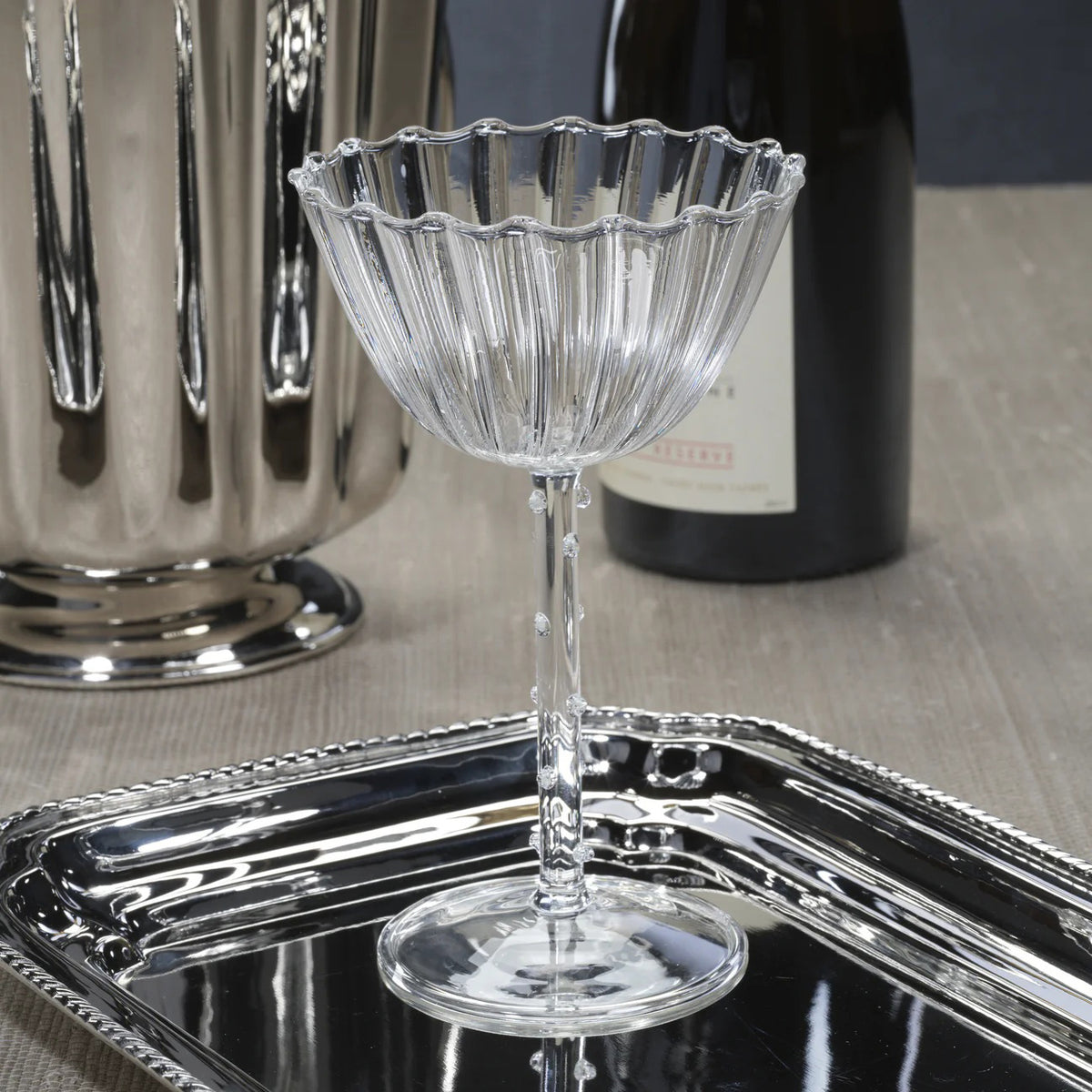 The ZODAX CHAUMONT Coupe Glass (260 ml), crafted from clear fluted Borosilicate glass, sits on a reflective silver tray with a metallic vase and wine bottle softly blurred in the background against a neutral surface.