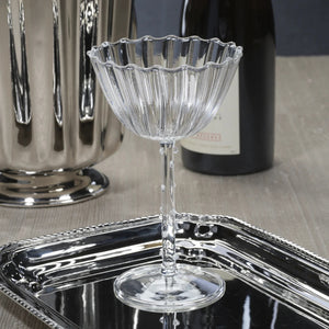 The ZODAX CHAUMONT Coupe Glass (260 ml), crafted from clear fluted Borosilicate glass, sits on a reflective silver tray with a metallic vase and wine bottle softly blurred in the background against a neutral surface.