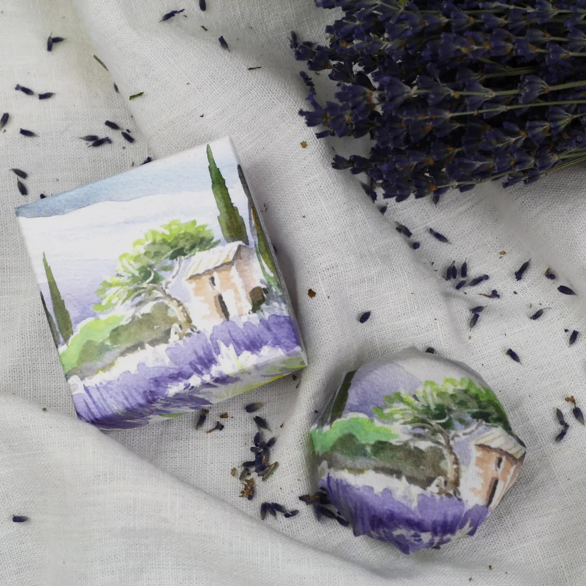 L&#39;AUGUSTE PROVENCE&#39;s THE ESSENCE OF PROVENCE Lavender Soap with Gift Box, featuring handmade French soap and organic lavender soap in illustrated wrapping, is displayed on white fabric with dried lavender and a lavender bouquet nearby.