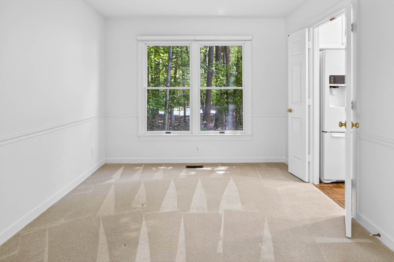 A bright, empty room with beige carpet, white walls, and a large window overlooking green trees. An open door reveals part of a kitchen with a refrigerator visible.