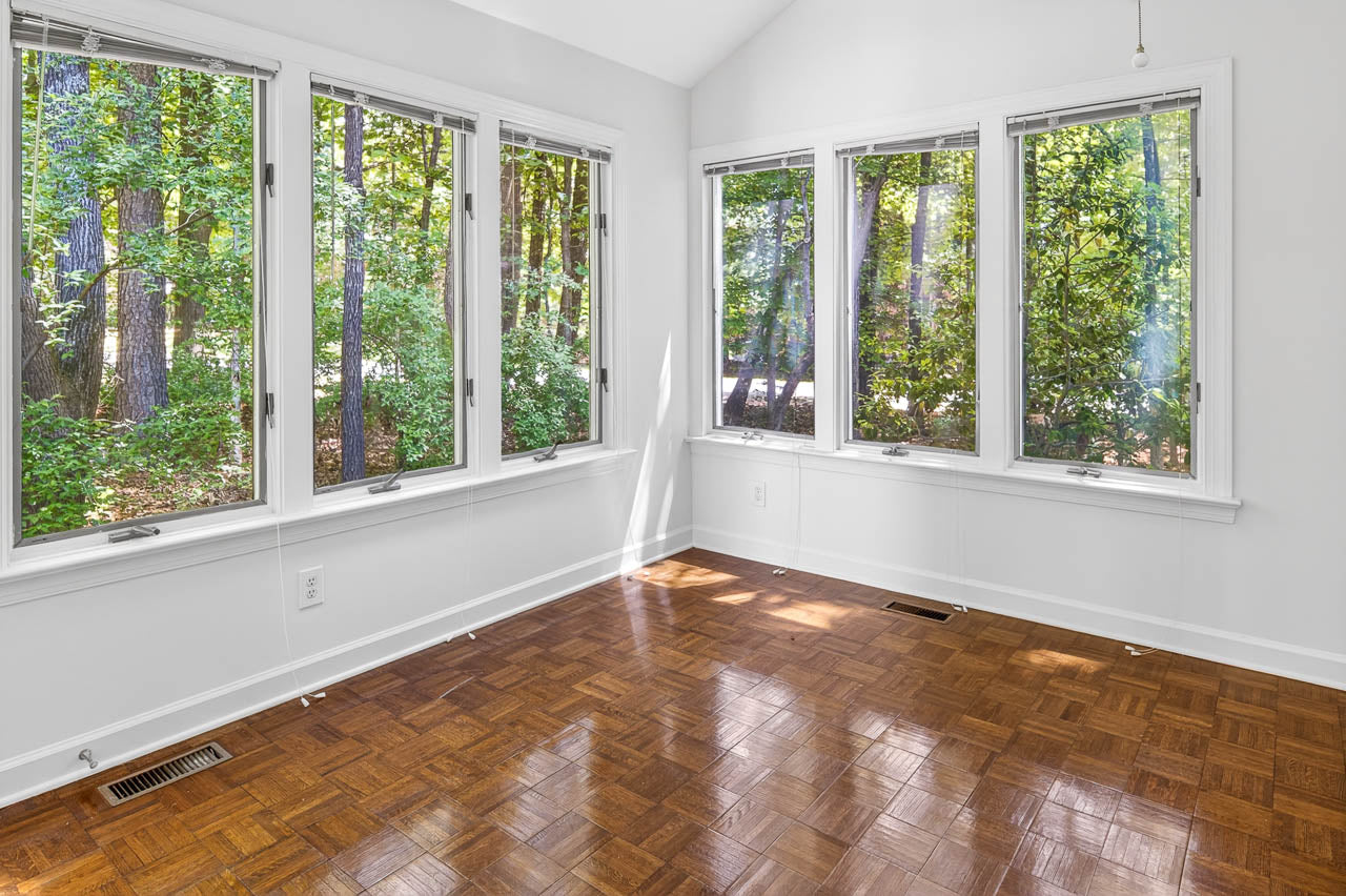 Bright sunroom with large windows, white walls, and a shiny wood parquet floor, overlooking a green, wooded area outside. Sunlight filters through the trees into the empty space.