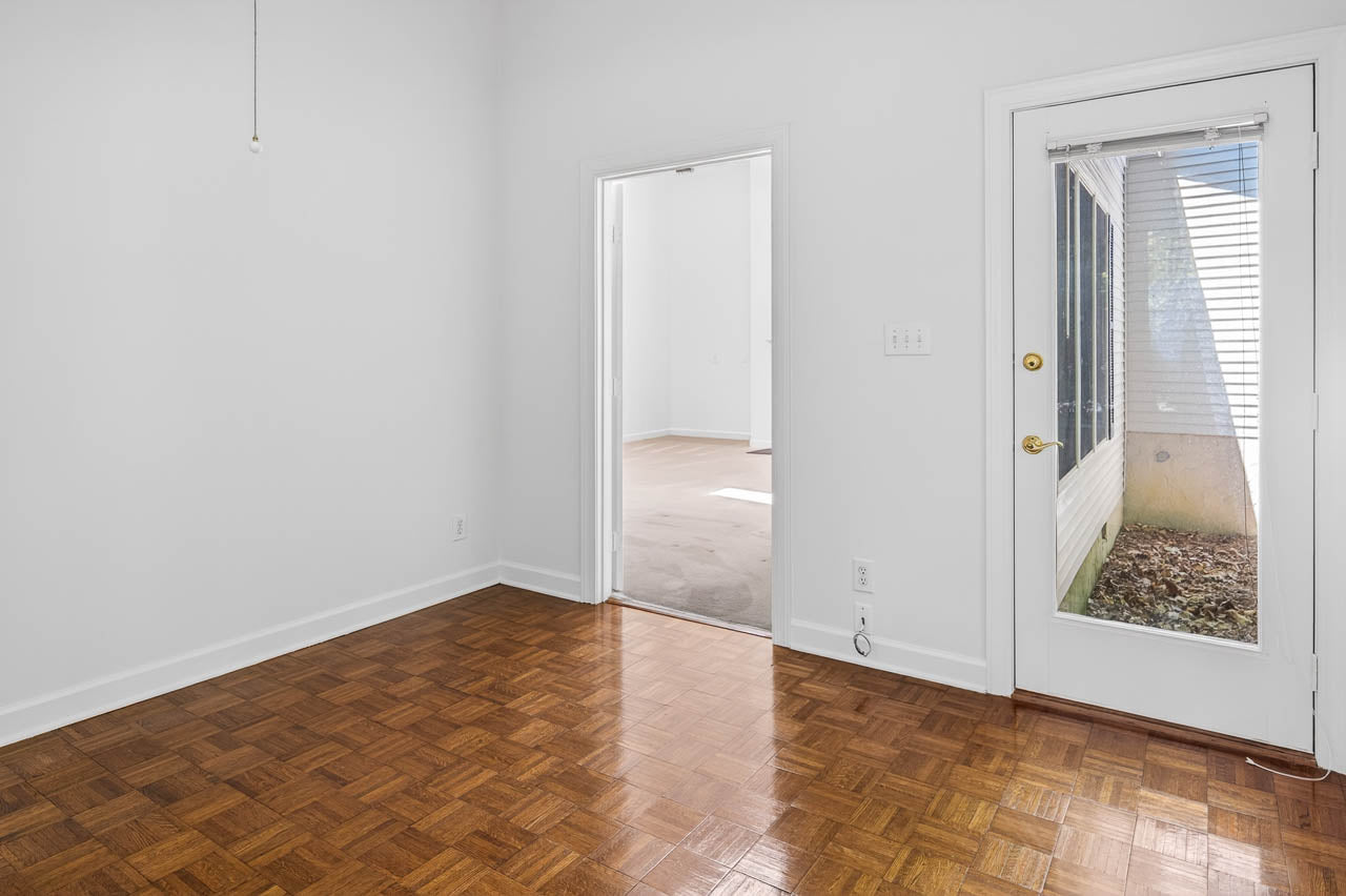 A bright empty room with parquet wood flooring, white walls, a glass door leading outside, and an open doorway connecting to another carpeted room. Sunlight filters through the blinds and glass door.