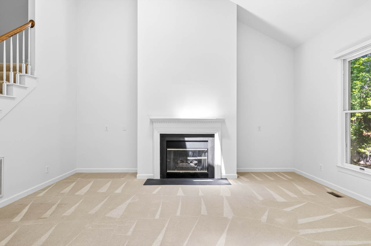 A bright, empty living room with clean beige carpet, a white fireplace centered on the far wall, high ceilings, and a large window letting in natural light. Stairs with a wooden railing are visible on the left.