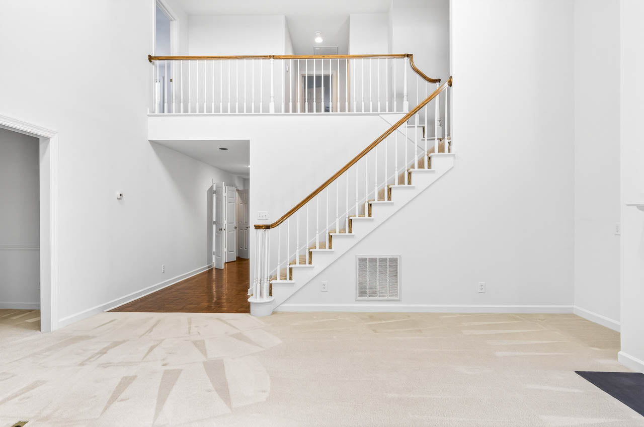 A bright, spacious interior with white walls, a wooden staircase with white railings leading to an upper floor, and light-colored carpet and hardwood flooring. Multiple doorways and a high ceiling are visible.