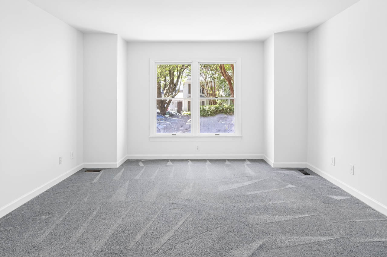 An empty, bright room with white walls, clean gray carpet, and a large window letting in natural light. Trees and greenery are visible outside through the window.