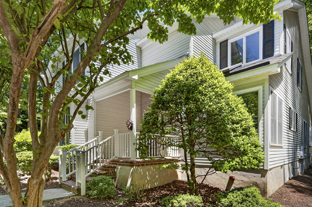 A white house with blue shutters features a covered front porch, brick steps, landscaped bushes, and a tree in the foreground providing shade.
