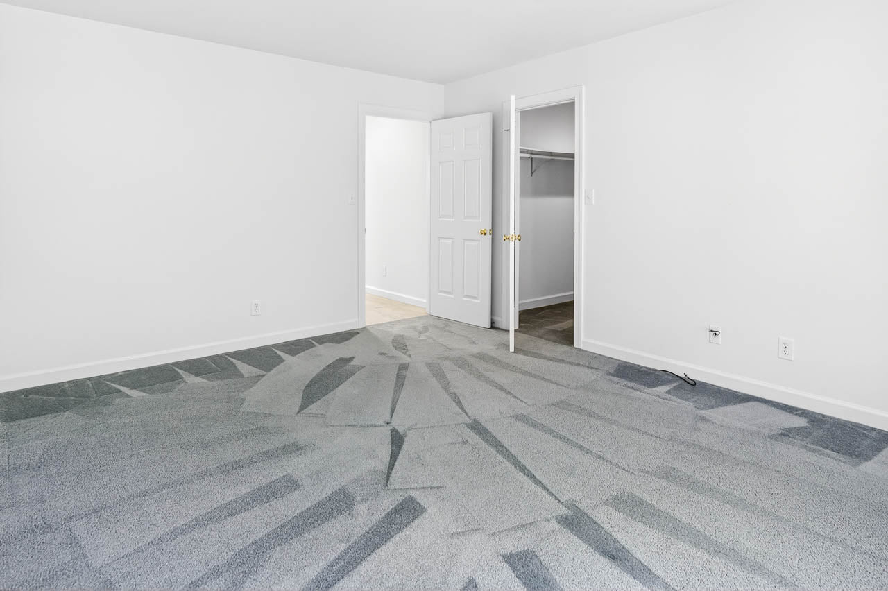 Empty room with freshly vacuumed light gray carpet showing patterns, white walls, two open white doors—one leading to a closet and the other to a hallway. Minimal electrical outlets and baseboards are visible.