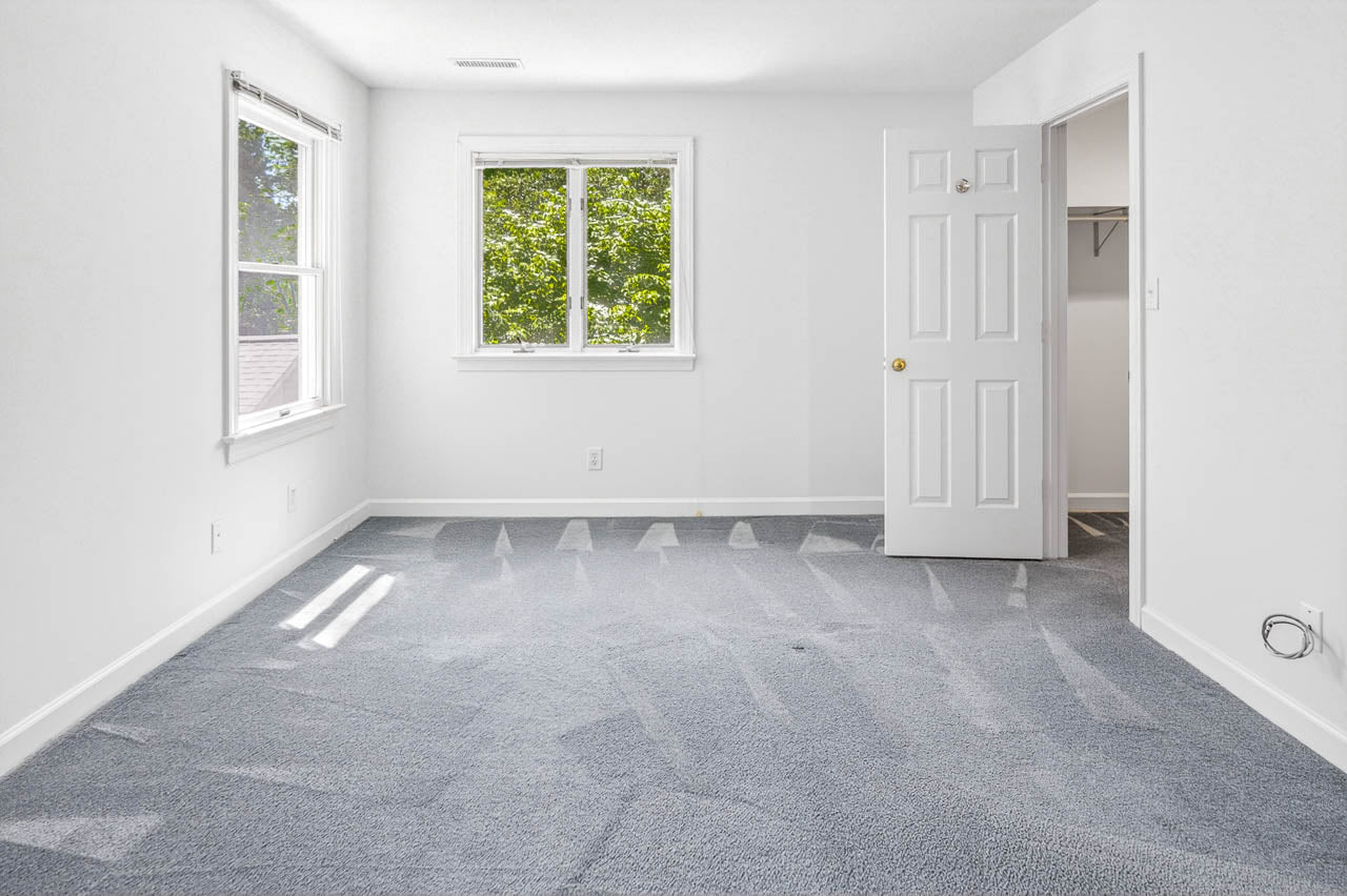 A bright, empty room with clean, light-gray carpet, two windows letting in natural light, white walls, an open door leading to a closet, and a few visible electrical outlets.