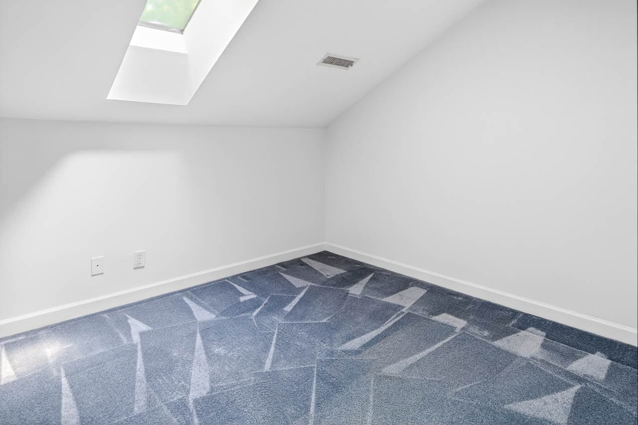 A small, empty room with white walls, a sloped ceiling, a skylight, and blue carpet with visible vacuum lines. There are electrical outlets and an air vent on the wall. Light streams in from the skylight.