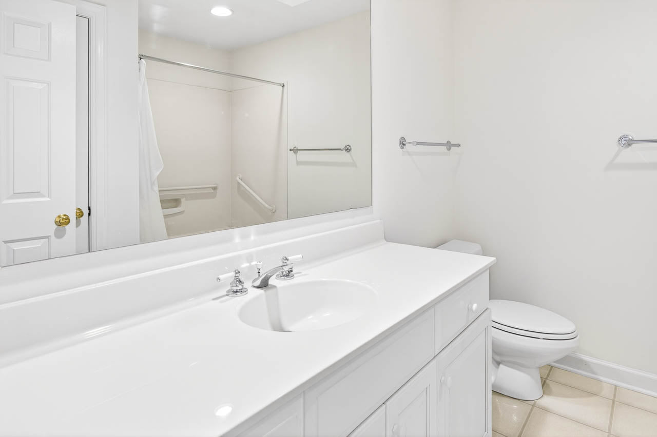 Bright, clean bathroom with white countertop, sink, and cabinetry. There’s a toilet and a shower with a glass door and grab bars. The room has tiled floors and a large wall mirror above the sink.