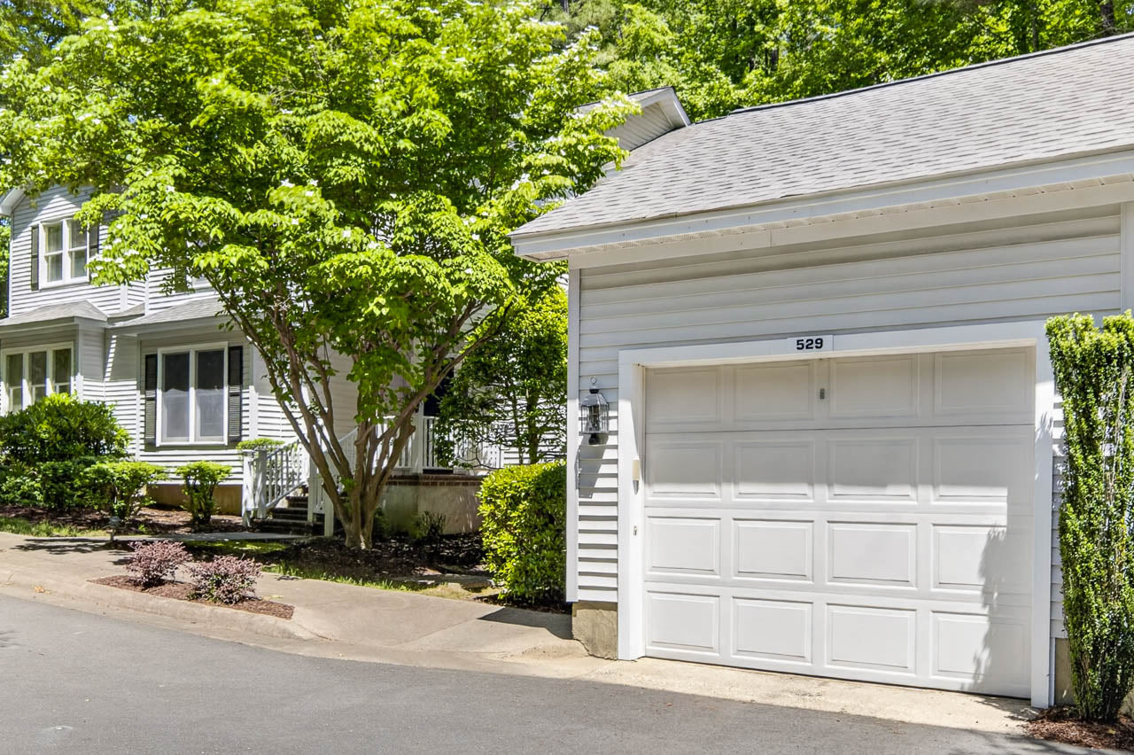 A white garage with the number 529 and a closed door is attached to a house. A lush tree and green shrubs are next to the building, and sunlight filters through the leaves onto the driveway and lawn.