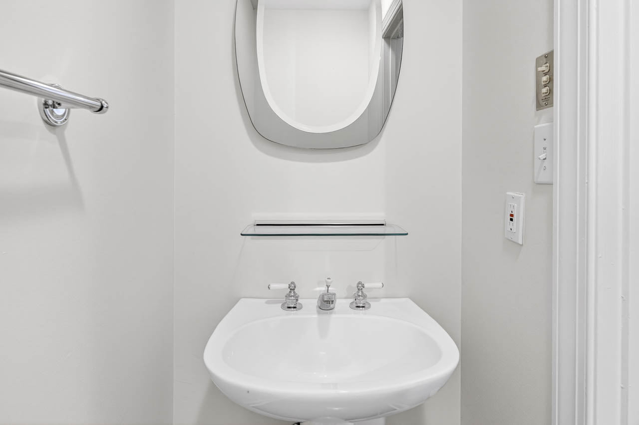 A small, white bathroom sink with silver faucets is mounted below a glass shelf and a modern, curved mirror on a plain white wall. A towel bar and light switches are visible on the left and right sides.