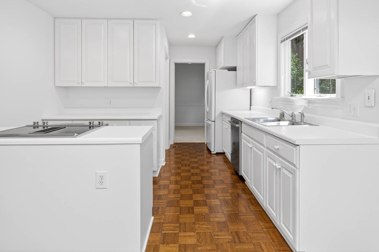 Bright, modern kitchen with white cabinets, countertops, and appliances, parquet wood flooring, a window above the sink, and recessed ceiling lights. A doorway leads to another room in the background.