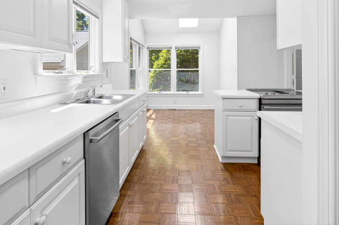 A bright, modern kitchen with white cabinets, stainless steel appliances, and parquet wood flooring, featuring large windows that let in natural light and offer a view of greenery outside.