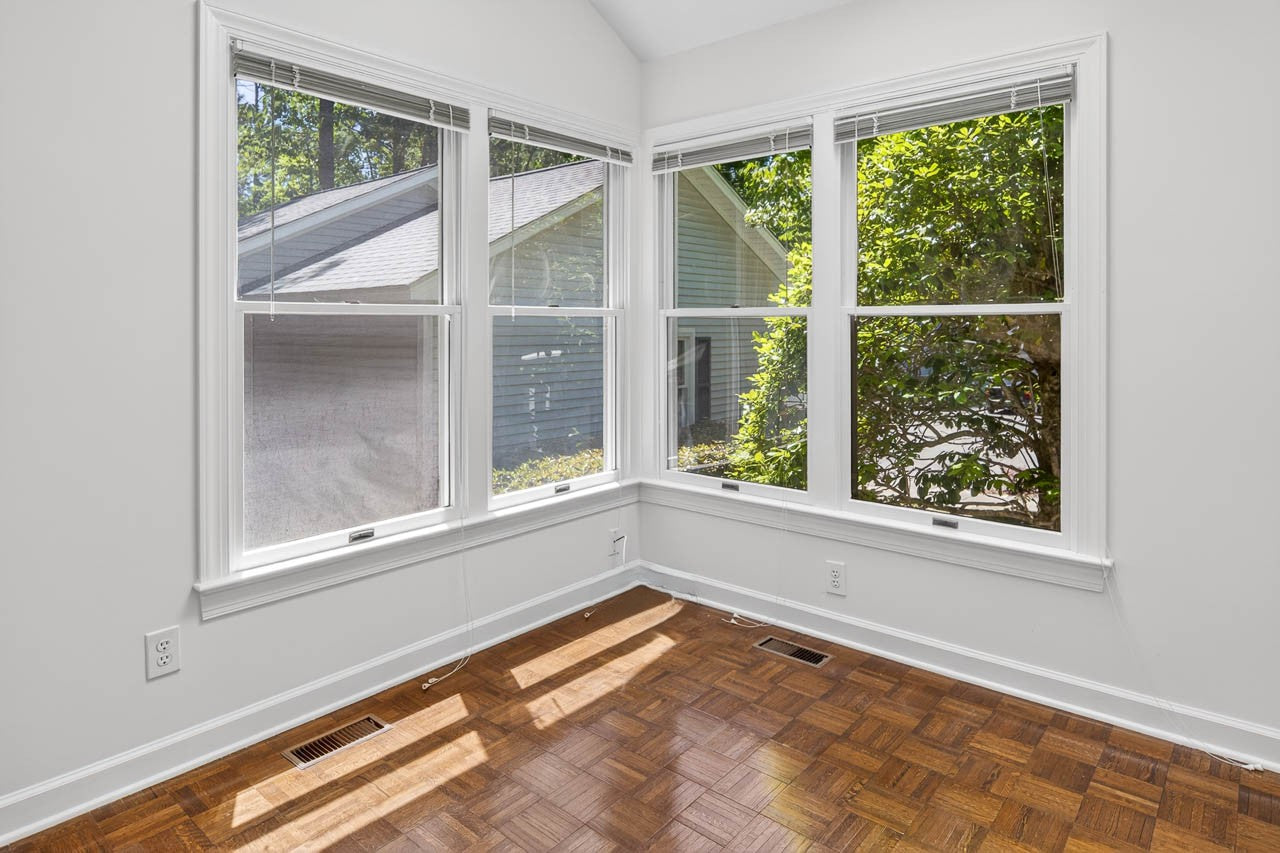 A bright corner of a room with large windows, white walls, and wooden parquet flooring. Sunlight streams in, and green trees and a neighboring house are visible outside.