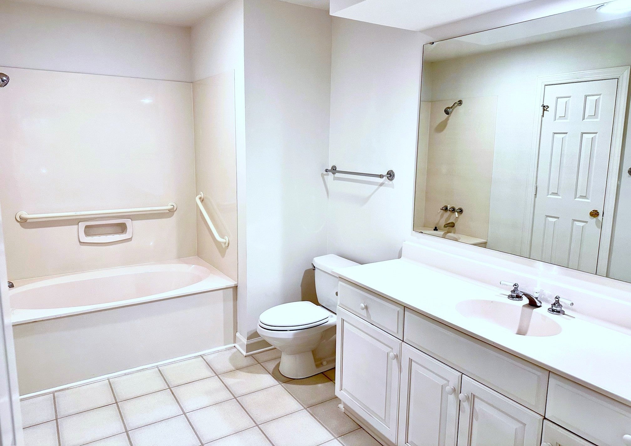 Accessible bathroom with a bathtub featuring grab bars, a toilet, and a long white vanity with a sink and large mirror. The walls and tiles are light-colored, creating a clean and bright appearance.