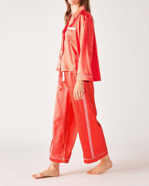 A woman models the MERSEA - OVER THE MOON SATIN SAILOR PAJAMA SET in teal with white trim, featuring a long-sleeved button-up top and elastic waist bottoms. She stands barefoot with her hands in the pockets, highlighting the silky smooth fabric.