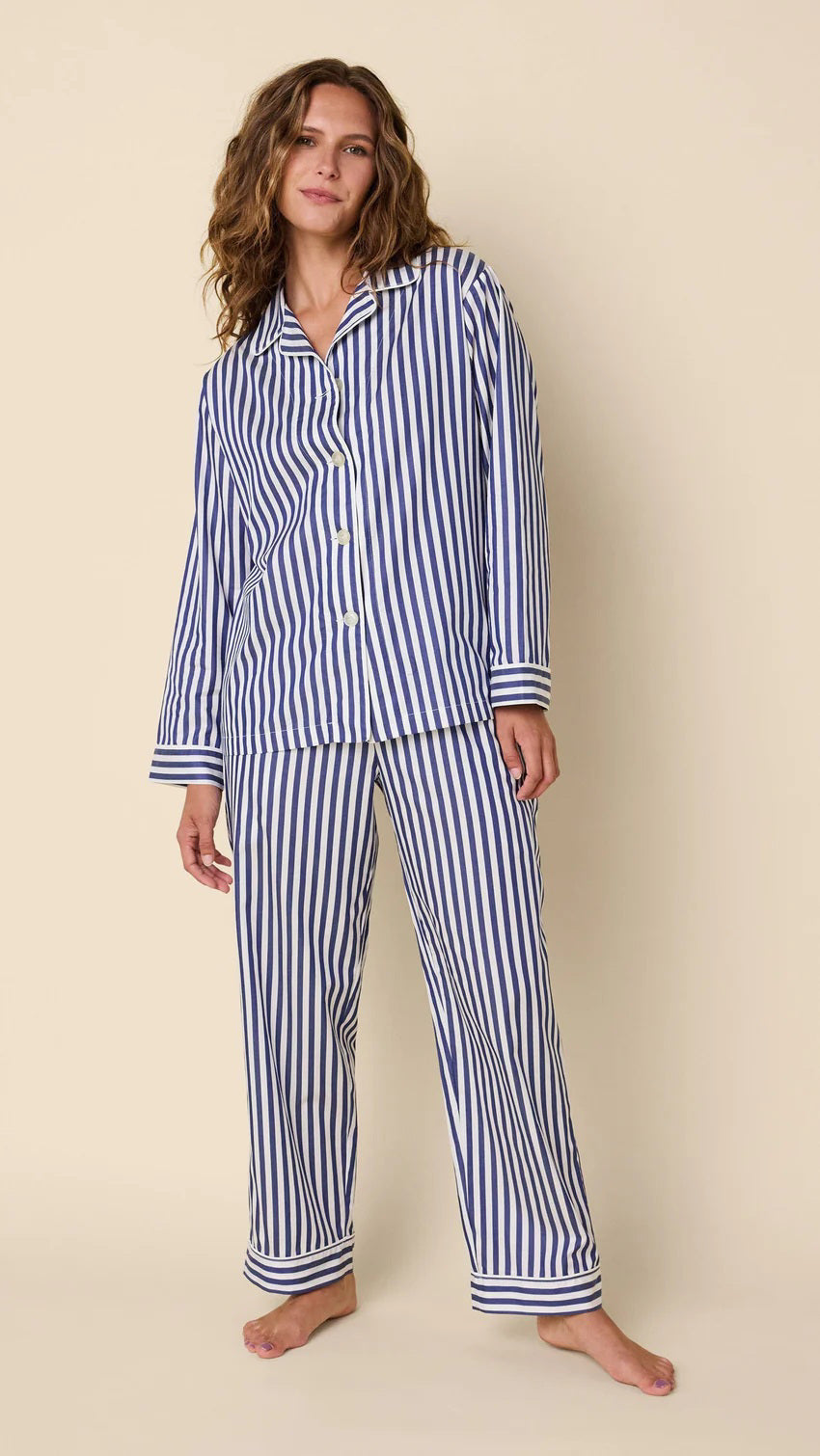A person with long wavy hair smiles while wearing the CAT&#39;S PAJAMAS Classic Stripe Long Sleeve pajama set. The background is plain light beige.