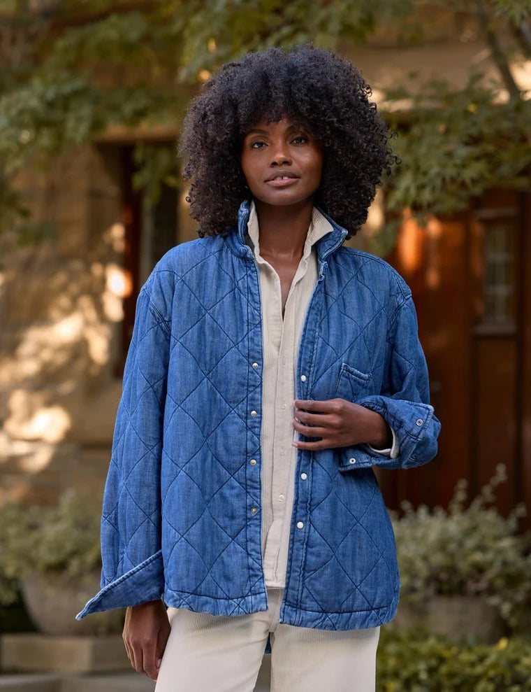 A person holds open a blue FRANK &amp; EILEEN - EILEEN Quilted Shirt Jacket in Famous Denim Distressed Indigo Wash, revealing its interior pocket and distinctive diamond quilted pattern. The person is wearing a cream-colored, ribbed top, with a blurred background.