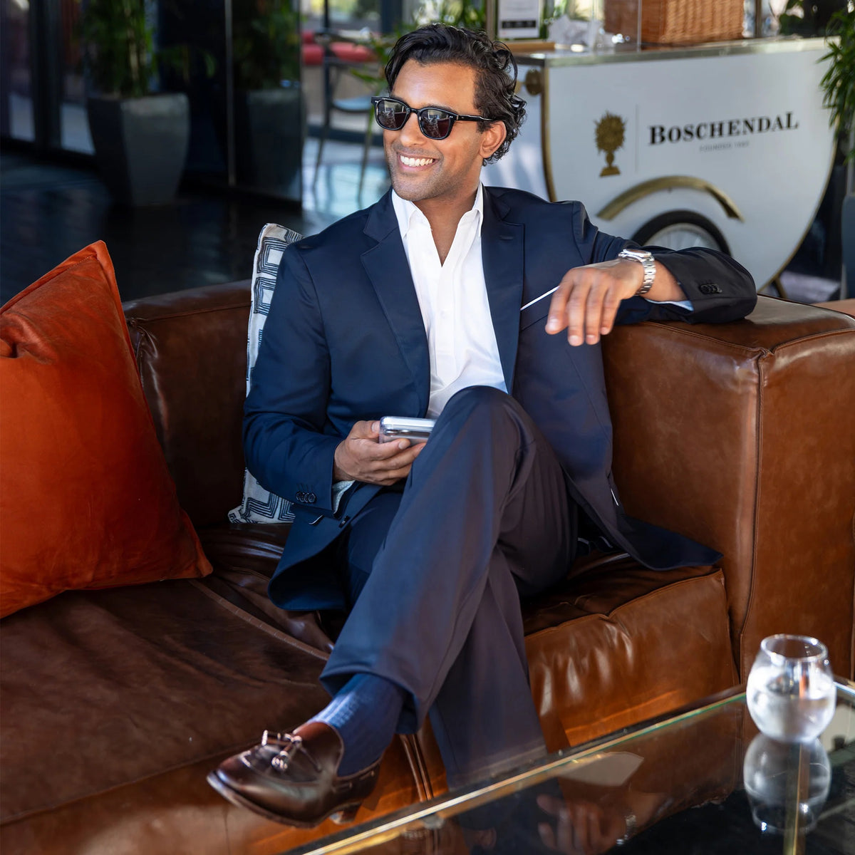 A man in COLLARS AND CO Maverick Performance Pants in Navy, a dark shirt, and sunglasses stands confidently outside a restaurant with green chairs and lush plants nearby, holding his phone.