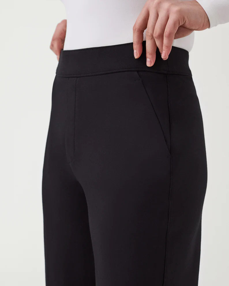 A woman stands against a plain background wearing the SPANX Perfect Pant Wide Leg in black—crafted from premium ponte fabric—paired with a white long-sleeve V-neck top and black heeled shoes.