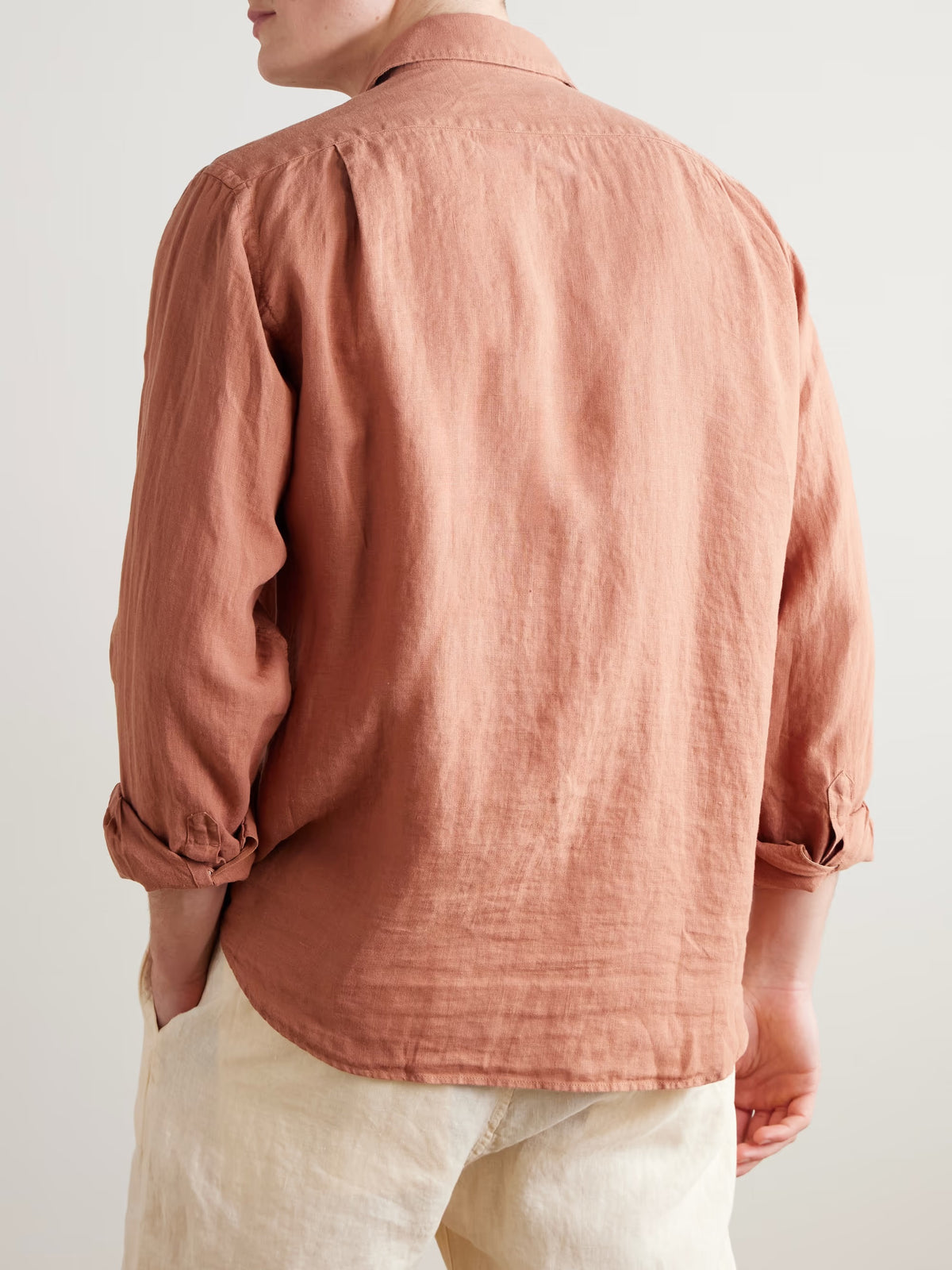 A person wears the HARTFORD - PAUL LINEN SHIRT IN FADED COPPER by HARTFORD, paired with light pants, standing with one hand in their pocket against a plain background.