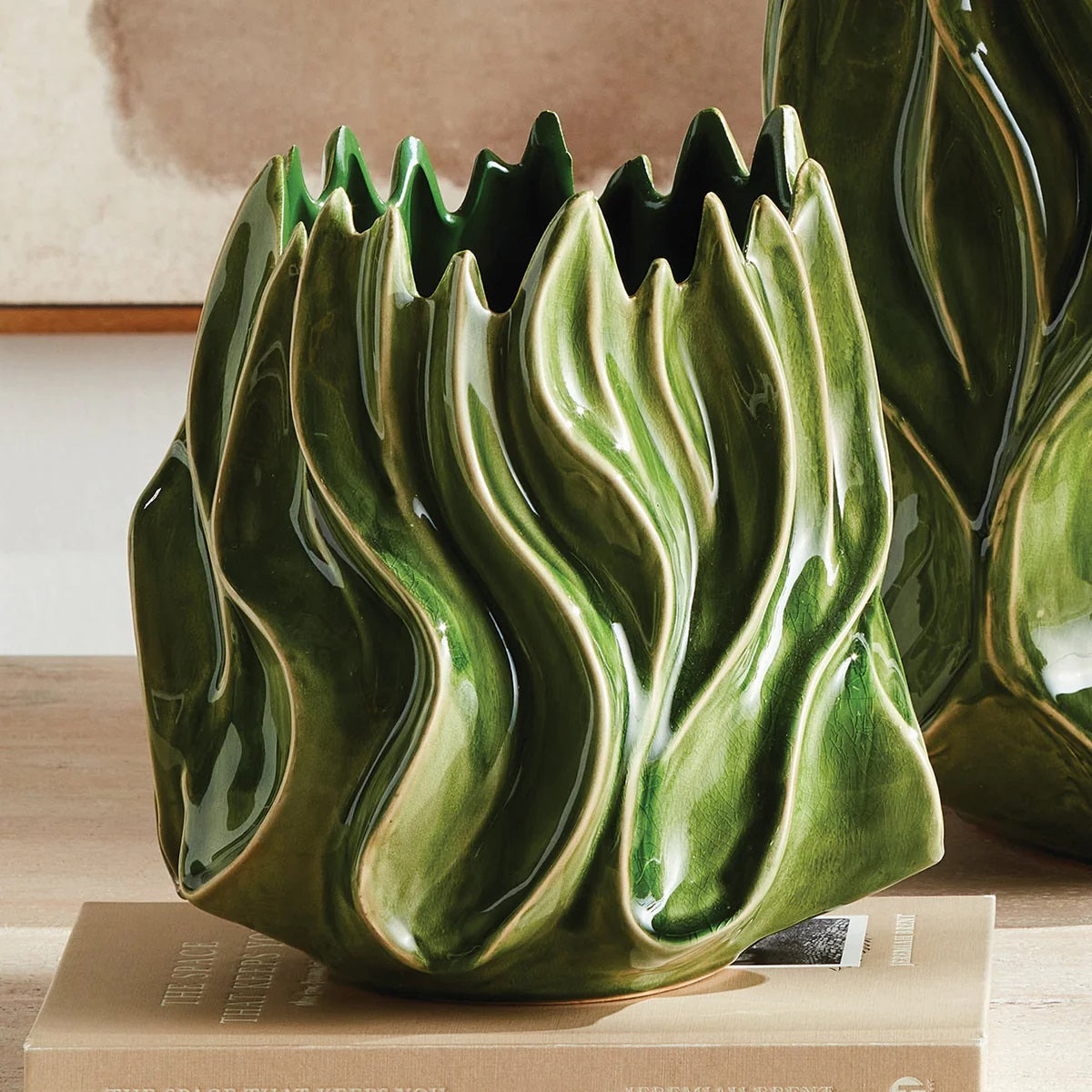The NAPA HOME AND GARDEN - GREEN CALLISTA VASE, MEDIUM features a sculptural design with glossy green handcrafted ceramic, showcasing wavy, flame-like edges and ridges that evoke the look of abstract leaves or flames. A high-gloss reactive glaze accentuates its flowing, organic exterior shapes, while the interior is finished in a rich dark green.