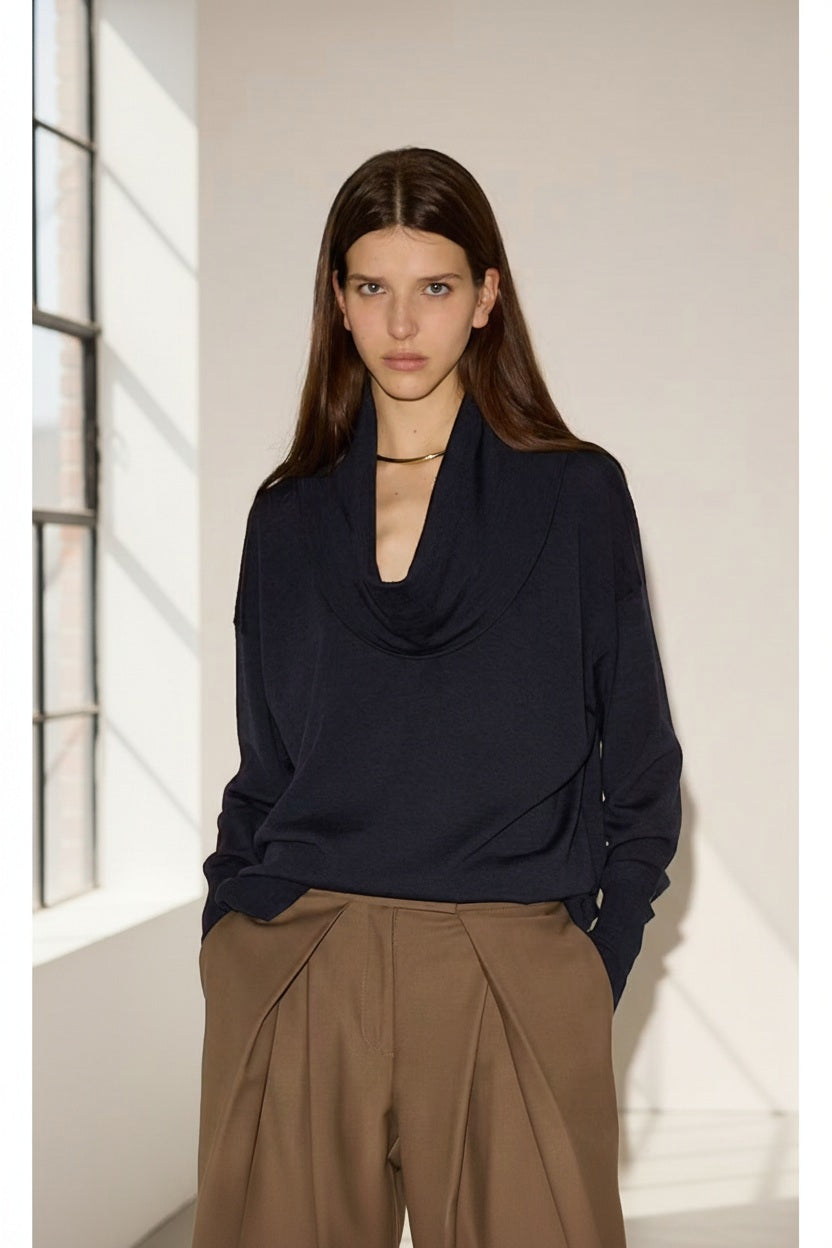 A woman with long brown hair stands indoors by a window, wearing the NUDE Draped Cowl Neck Sweater in blue virgin wool paired with high-waisted brown trousers. With her hands in her pockets, she looks directly at the camera, embodying effortless sophistication. This elegant piece from NUDE is made in Italy.