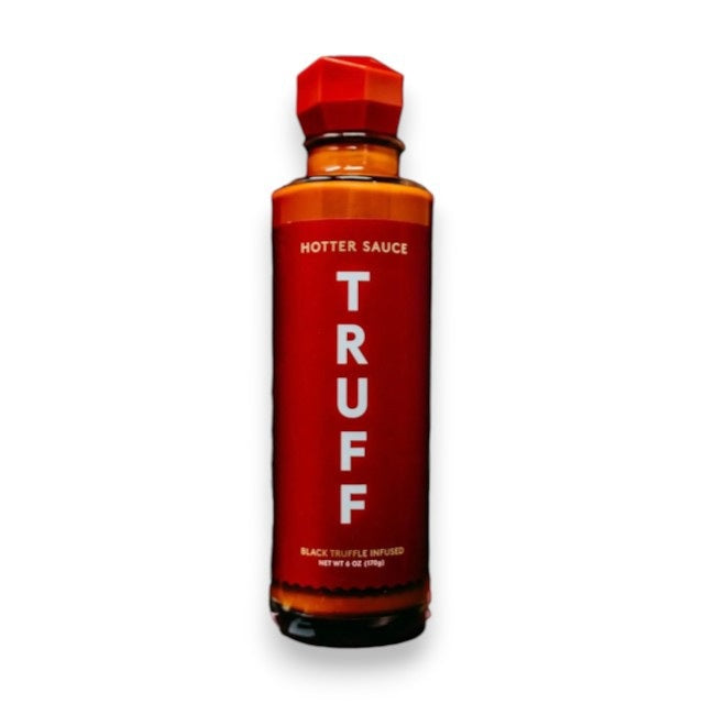 A bottle of TRUFF - HABANERO HOT SAUCE by TRUFF HOT SAUCE is being poured over a dish of macaroni and cheese in a white rectangular bowl, set against a brightly lit, white background.
