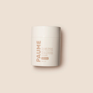 A white cylindrical container of PAUME’s OVERNIGHT HAND AND FOOT HYDRATION MASK rests on a creamy textured surface, with nourishing mask—ideal for dry, cracked skin—smeared around it.