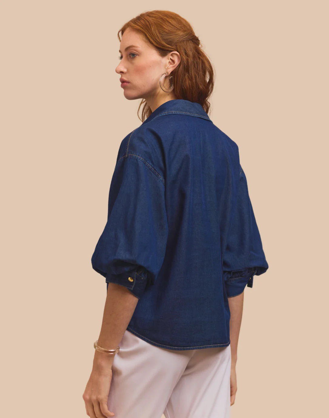 A woman with red hair faces away, wearing the HINSON WU Lilian Denim Elbow Sleeve Blouse in dark blue and white pants, set against a beige background.