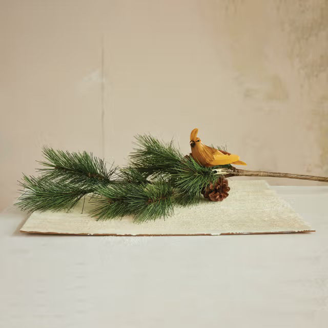 A pine branch with needles and a pine cone rests on a textured white surface. The CREATIVE COOP CARDINAL CLIP ON ORNAMENT is attached to the branch, set against a beige background.