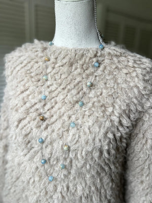 A mannequin in a beige textured sweater showcases the ANN LIGHTFOOT - BLUE CALCITE INTERMITTENT STRAND NECKLACE, a delicate 34" piece with spaced blue and brown beads on a clear strand. The softly blurred background highlights the jewelry.
