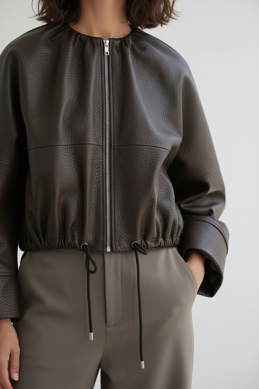 A person wearing the MEIMEIJ FAUX LEATHER BOMBER JACKET in dark brown faux leather with a textured finish and adjustable drawstring hem, paired with taupe high-waisted pants, stands against a plain light background. The jacket by MEIMEIJ lends a modern edge to the outfit, while the person’s head is partially out of frame.