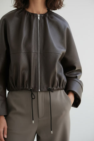 A person wearing the MEIMEIJ FAUX LEATHER BOMBER JACKET in dark brown faux leather with a textured finish and adjustable drawstring hem, paired with taupe high-waisted pants, stands against a plain light background. The jacket by MEIMEIJ lends a modern edge to the outfit, while the person’s head is partially out of frame.