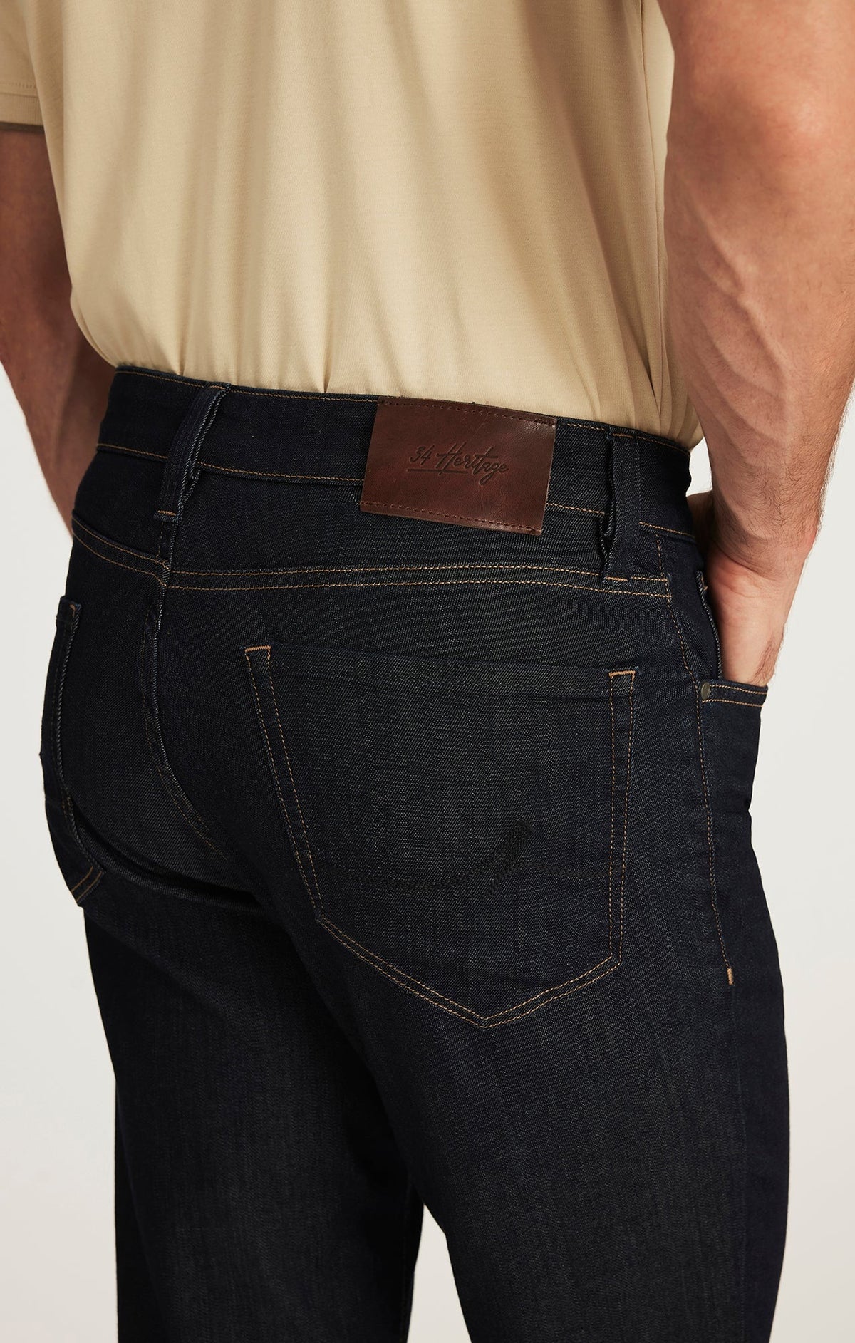 A man wearing a beige short-sleeve shirt, tan suede shoes, and the 34 HERITAGE - CHARISMA RELAXED STRAIGHT JEANS IN RINSE VINTAGE by 34 HERITAGE stands with one hand in his pocket against a white background.