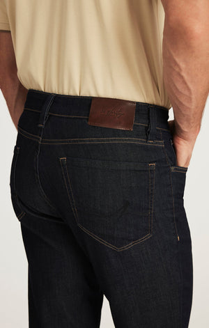 A man wearing a beige short-sleeve shirt, tan suede shoes, and the 34 HERITAGE - CHARISMA RELAXED STRAIGHT JEANS IN RINSE VINTAGE by 34 HERITAGE stands with one hand in his pocket against a white background.