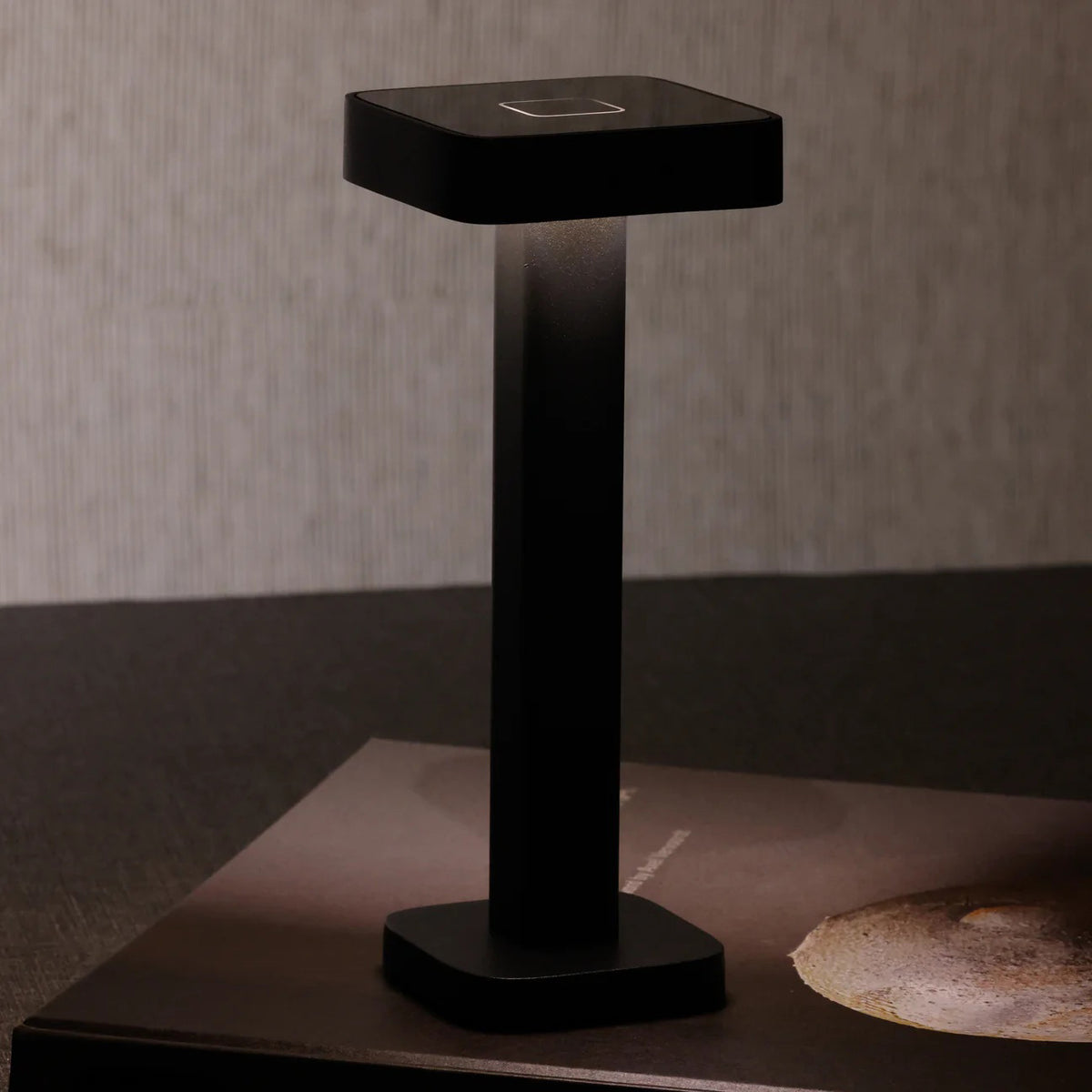 The ZODAX MONZA RECHARGEABLE TABLE LIGHT is a black, rectangular, pedestal-shaped device with a square touchpad on top, offering USB rechargeable capability and adjustable light intensity, shown upright on a white background.