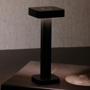 The ZODAX MONZA RECHARGEABLE TABLE LIGHT is a black, rectangular, pedestal-shaped device with a square touchpad on top, offering USB rechargeable capability and adjustable light intensity, shown upright on a white background.