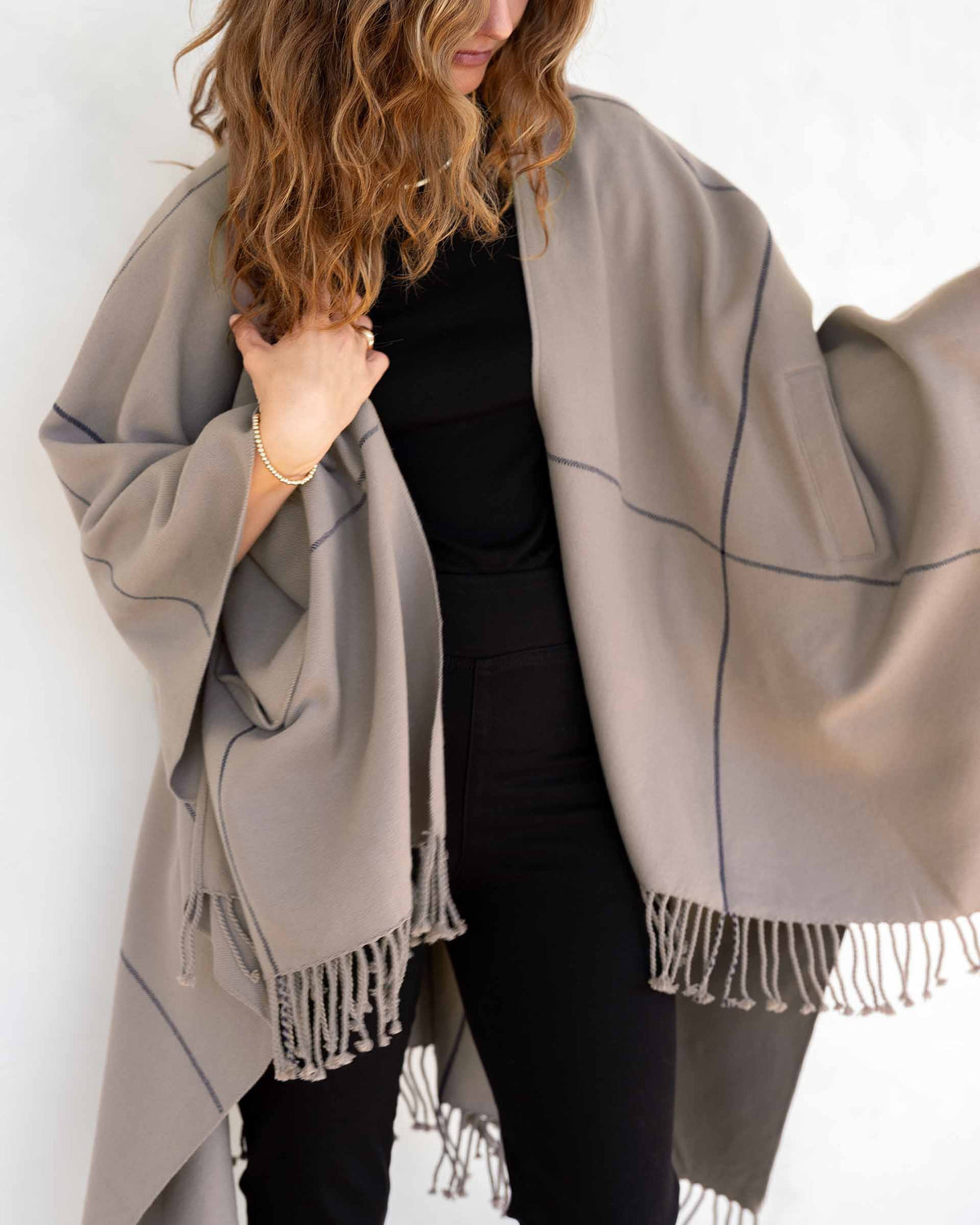 A woman with wavy brown hair wears the MERSEA Plaid Pocket Wrap—a black, white, and red plaid poncho with fringed edges—over a white outfit indoors. This MERSEA travel wrap is perfect for cozy style on the go.