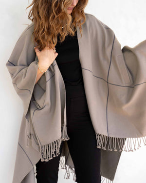 A woman with wavy brown hair wears the MERSEA Plaid Pocket Wrap—a black, white, and red plaid poncho with fringed edges—over a white outfit indoors. This MERSEA travel wrap is perfect for cozy style on the go.