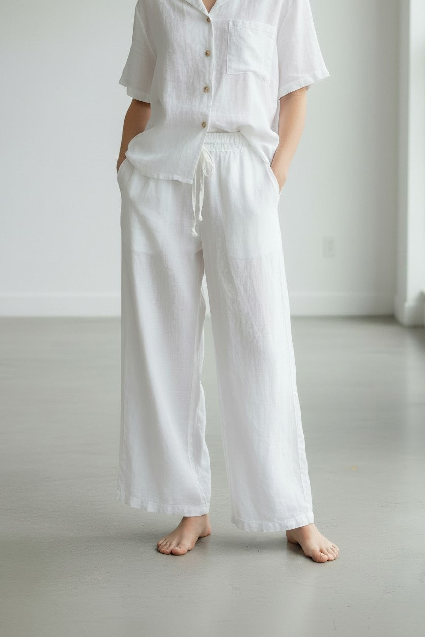 A person stands barefoot on a light floor, wearing the LLEA BLUE - DANA Straight Leg Drawstring Linen Pant in White by LLEA BLUE, styled with a loose silhouette. They pair the pants with a white short-sleeve button-up shirt featuring a chest pocket. The image is cropped at the shoulders, set against a minimalistic background.