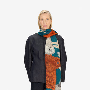 A woman faces forward, wearing a dark jacket and pants, black shoes, and the INOUI EDITIONS TOUTOU DOGS wool scarf in duck blue—one of our unique, hand-printed washable wool scarves from INOUI.