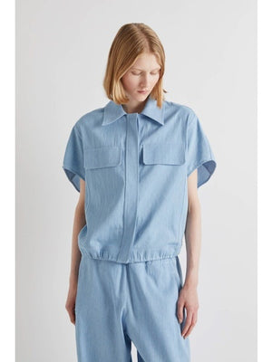 A person with chin-length light brown hair stands with their back to the camera, wearing the MEIMEIJ DRAWSTRING COTTON TOP in chambray blue. The loose fit and plain white background highlight MEIMEIJ's modern Italian aesthetic.