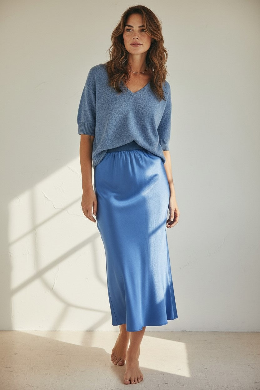 A woman with wavy brown hair stands barefoot indoors in a light blue V-neck short-sleeve sweater and the ROSE & CROWN - SATIN MIDI SKIRT by ROSE & CROWN, featuring a silky blue satin finish and elastic waistband, against a plain light wall.